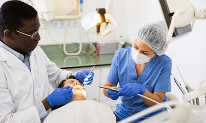 Professional african american dentist performing teeth treatment to woman with help of female assistant in dental clinic..