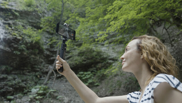 Woman taking pictures or shooting video of mountains and waterfall on her smartphone using a tripod. Action. Female traveler with a selfie stick and a phone standing in a gorge surrounded by green