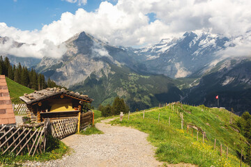 Fototapeta premium Beautiful Swiss mountains in white clouds, snow on the peaks and green meadows, a sandy stone path along a wooden house with a fence to the red national flag