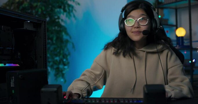 Young Asian woman is playing video game on computer at night while talking online through headset with team, women is making head-to-toe body movements while avoiding obstacles in virtual world
