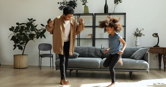 Little Black girl teach young adult mommy older sister nanny to dance disco feel music rhythm at cozy living room. Smiling millennial mom try to repeat dance moves after tween daughter on warm floor