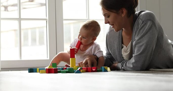 Caring mom lie on warm floor at light living room build house from bright colorful wooden brick constructor set with cute infant child. Little baby learn to play simple toys together with mother nanny