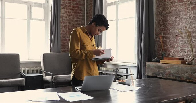 Busy young Afro American woman stand by desk involved in work with paper and electronic documents make notes in daybook. Millennial female freelancer analyze marketing reports at home office workplace