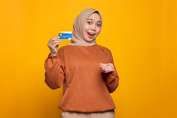Shocked young Asian woman in orange shirt holding money banknotes and showing credit card isolated over yellow background