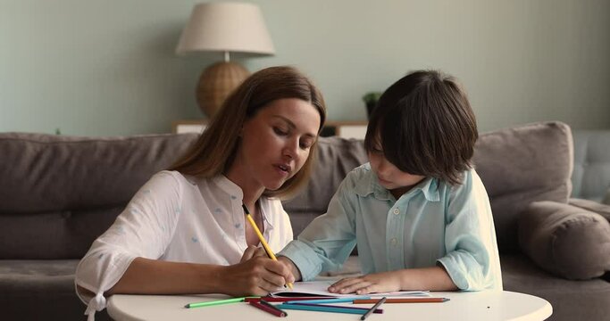 Young beautiful woman her preschool 6s son drawing pictures in sketchbook, sit at table in modern living room talk enjoy favourite hobby and carefree weekend leisure. Kid development, pastime concept