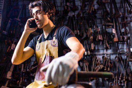 Authentic blacksmith man wear leather apron in dark indoors studio calling by phone - Powered by Adobe