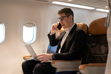 Cheerful businessman in airliner using laptop computer while talking on cellphone.