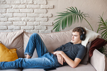 teenage boy with dark hair in a gray T-shirt and jeans with headphones lying on the couch at home. boy listens to music or a podcast, the smartphone is lying nearby. Take a break from your eye phone
