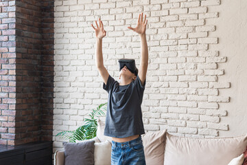 A teenage boy wearing glasses in a VR headset plays virtual games in his room. The young man raised his hands. Entertainment of modern teenagers using technology.