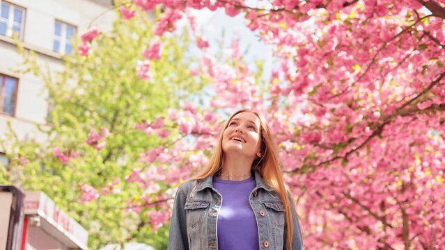 Portrait of beautiful young woman look around and smiling on blurred background in the city street. Sakura tree. Happy Caucasian beautiful female walking around Cherry blossom trees. Slow motion.