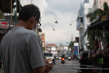 young asian man standing playing smartphone, gray shirt, glasses, defocused background