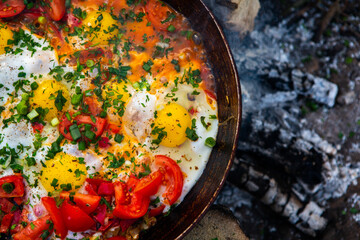 Eggs prepared with vegetables on a frying pan on the fire in the forest. Cooking scrambled eggs over an open fire. Breakfast in nature. Tourist food.