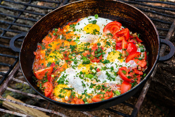 Eggs prepared with vegetables on a frying pan on the fire in the forest. Cooking scrambled eggs over an open fire. Breakfast in nature. Tourist food.