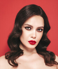 Fototapeta premium Beauty portrait of young brunette woman with evening makeup and perfect clean skin. Sexy model with curly hair posing in studio. With red bright natural lips. Isolated on red background