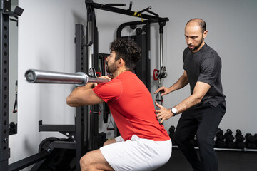 Fitness instructor giving support to young man lifting barbell at health club