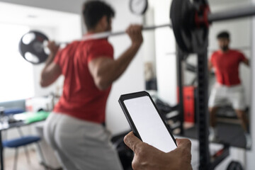 Hand of coach holding smart phone near young man lifting barbell at gym