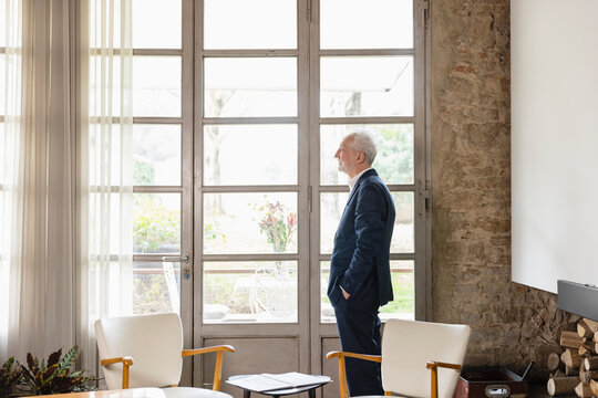 Man with hand in pocket looking through widow standing in living room at home