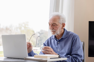 Senior man with laptop and coffee cup sitting by window at hotel apartment