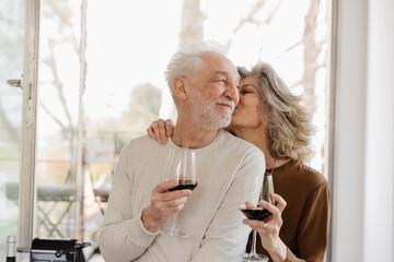 Senior woman holding wineglass kissing man in front of window at hotel apartment