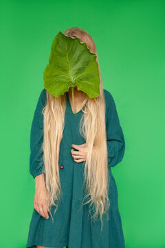 Blond woman covering face with burdock leaf against green background