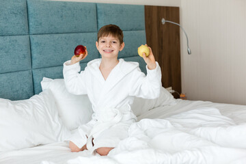 A child in a white coat on the bed is smiling and holding apples. Health. Morning.
