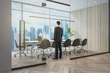 Attractive european businessman standing in modern concrete and wooden, glass conference room interior with window and blurry city view, furniture and equipment.