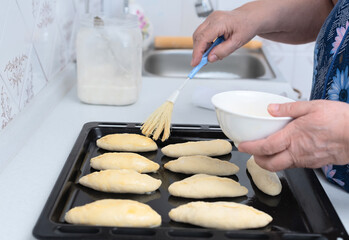 Senior woman hands brushing raw pies with egg before baking on a baking tray. Selective focus. Process of making pies with apple filling. Cooking at home concept. Tradition home-made food