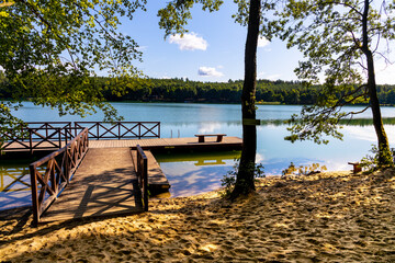 Wooded shore of Jezioro Gwiazdy lake with wooden jetty platform in Bukowo Borowy Mlyn Village near Bytow of Pomerania in Kashubian region of northern Poland - obrazy, fototapety, plakaty