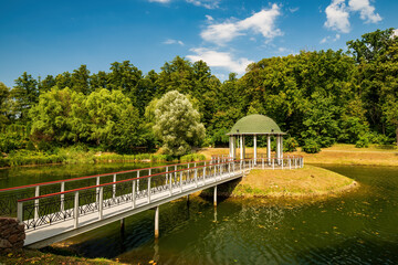 Fototapeta na wymiar Small island in the center of park pond with round gazebo and pedestrian bridge. Feofaniya public park, Kyiv, Ukraine