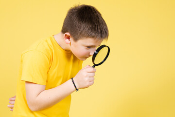 Cute boy playing to be a detective and searching clues using a loupe isolated on yellow background