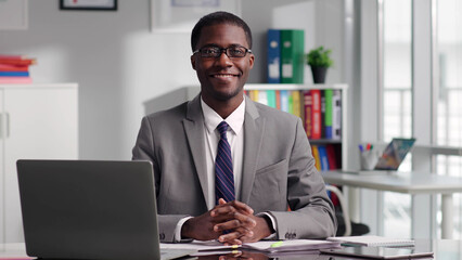 Portrait of African-American politician in glasses smiling at camera sitting at desk  - obrazy, fototapety, plakaty