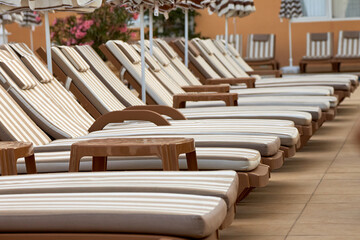 Plastic deck chairs with soft mattresses and umbrellas from the sun stand by pool for those who like to sunbathe. Sun loungers for relaxing and sunbathing are located in foreground next to the pool