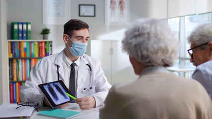 Young doctor in safety mask show x-ray on tablet to aged couple