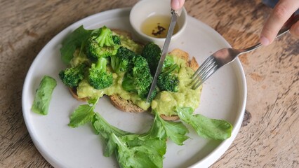 A healthy breakfast of toast with avocado and broccali cabbage sauce. A white plate of fried toast with avocado spread and green broccoli cut with a knife, dressing sauce and lettuce.