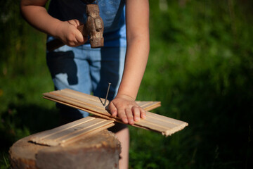 Child hardens nail into plank. Girl learns to beat with hammer. Preschooler is engaged in manual labor. Learning without computer. Work in fresh air.