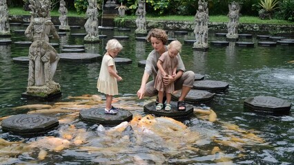 A young mother with two daughters in an Asian park feed the fish in the pond. A large flock of golden carps swam up to the children with their mother who feed them. Mother with children on the lake.