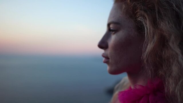 Abstract defocused shot of beautiful young caucasian woman with curly blond hair and freckles looking at camera. Cute woman portrait in a pink long dress posing on a volcanic rock high above the sea