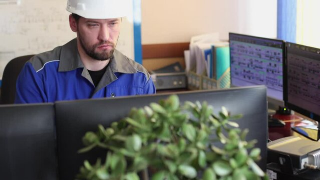 Serious Caucasian unshaven worker in overalls and helmet sits in the control room and uses a computer to monitor the correct operation of all systems. Automated workplace of the dispatcher or operator