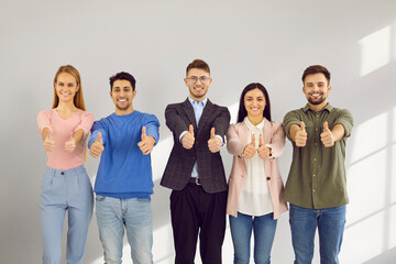 Portrait of active and smart young people showing thumbs up approving or recommending. Smiling diverse men and women casual clothes stand in row on gray wall background. Concept of positive approval