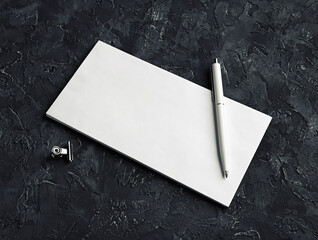 Photo of blank envelope, pen and metal clips on black plaster background. Blank stationery set.