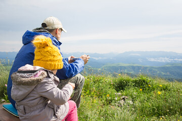 Fototapeta na wymiar family father and child girl in yellow hat sit and eat on background of nature - mountain landscape. travel with kids.