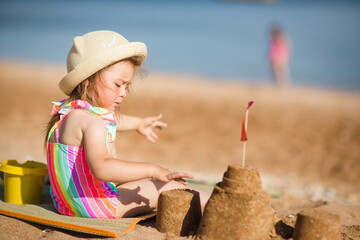 baby child in hat sits on beach against background of sea and plays sand, builds castle. vacation with kids in sunny summer