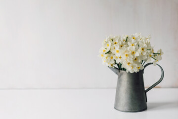 Bouquet of white daffodils in vintage vase on table or desk with space for text. Fresh spring flowers in metal jug. Minimal floral template. 