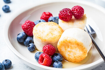 Cottage cheese pancakes with raspberries, blueberries and syrup in white bowl, gray background. Russian cuisine concept.