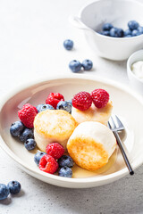 Cottage cheese pancakes with raspberries, blueberries and syrup in white bowl, gray background. Russian cuisine concept.
