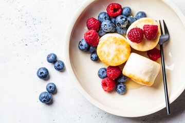 Cottage cheese pancakes with raspberries, blueberries and syrup in white bowl, gray background. Russian cuisine concept.