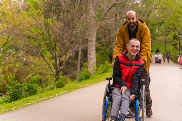 Paralyzed young man in the wheelchair being pushed by a friend in a public city park, strolling along a path having fun in spring
