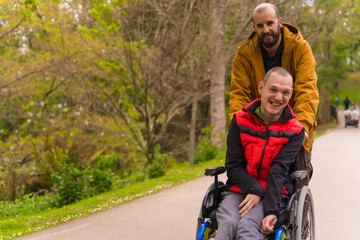 Portrait of a paralyzed young man in a wheelchair being pushed by a friend in a public city park,...