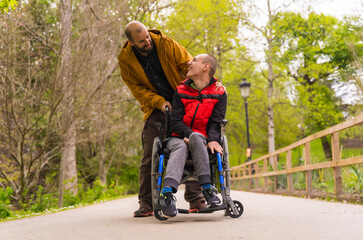 Paralyzed young man in the wheelchair being pushed by a friend in a public city park, strolling along a path having fun and smiling