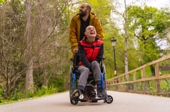 Paralyzed young man in the wheelchair being pushed by a friend in a public city park, strolling along a path having fun and smiling